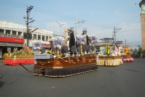 Ein Paradewagen mit Menschen darauf, umgeben von Gebäuden, Strommästen mit Drähten, Laternenmästen, Bäumen und einem Uhrturm im Hintergrund, unter einem bewölktem Himmel.