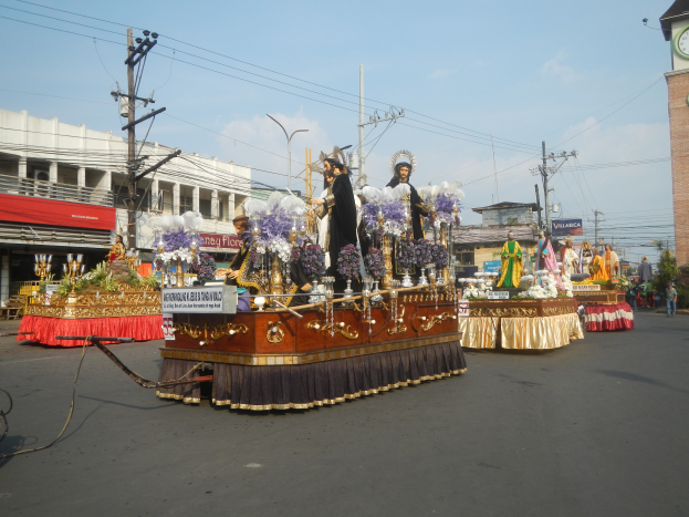 Ein Paradewagen mit Menschen darauf, umgeben von Gebäuden, Strommästen mit Drähten, Laternenmästen, Bäumen und einem Uhrturm im Hintergrund, unter einem bewölktem Himmel.