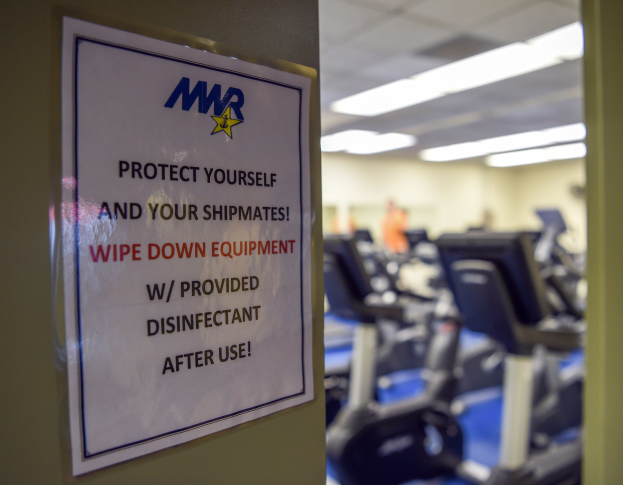 Ein Türschild mit der Aufschrift "Schützen Sie sich und Ihre Schiffskameraden" vor Laufbändern und Deckenlampen im Hintergrund, mit leichter Bewegungunschärfe.