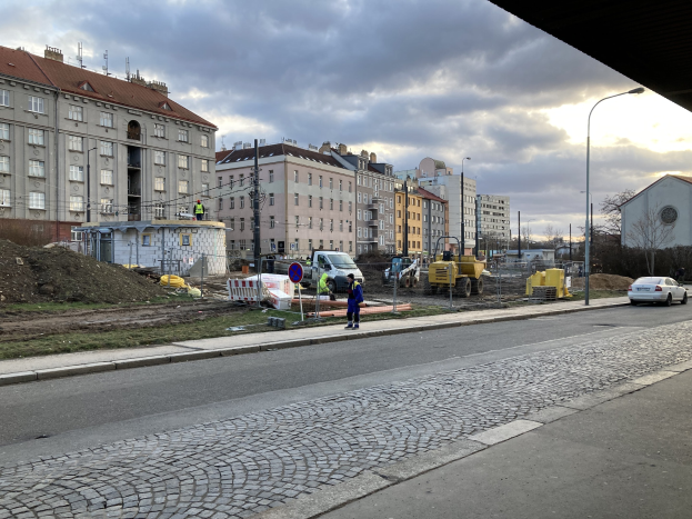 Stadtstraße mit parkenden Autos, Gebäuden, Laternen, Bäumen und einem Himmel mit Wolken, mit einer Straßensperre mit Fahrzeugen und Fußgängern im Vordergrund.