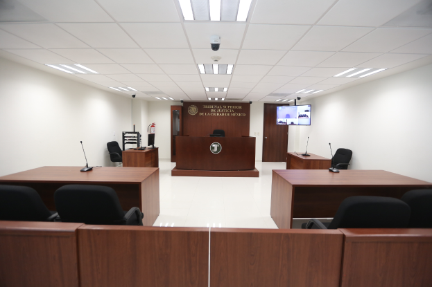 Ein Gerichtssaal mit hölzernen Tischen und Stühlen, Mikrophonen, einem großen Bildschirm, einer Tür, einer Texttafel, einem Feuerlöscher und Deckenleuchten.