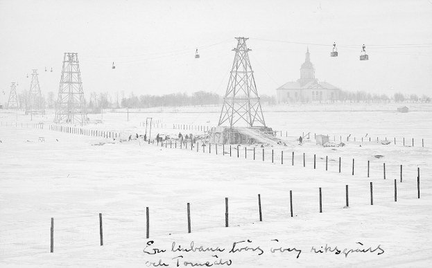Schwarze und weiße Fotografie einer Skihebe in einem verschneiten Feld mit Stützpfählen, Überseilbahn, Bäumen und einem Gebäude im Hintergrund, mit Text am unteren Rand.