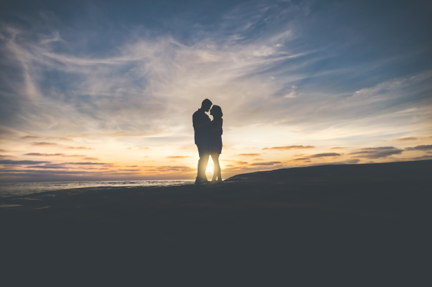Silhouette eines Paares, das sich auf einem Strand bei Sonnenuntergang umarmt und küsst, mit leuchtenden Orange-, Pink- und Purpurhimmel, tiefblauem Wasser und der untergehenden Sonne im Hintergrund.