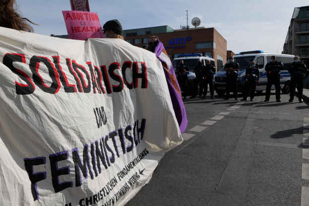 Eine Gruppe von Menschen geht eine Straße entlang und hält eine 'Solidarität und Feminismus'-Fahne, mit parkenden Fahrzeugen und Gebäuden im Hintergrund unter einem klaren blauen Himmel.