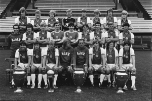 Ein Schwarz-Weiß-Foto einer grinsenden Fußballmannschaft, die mit ihren Pokalen posiert, einige sitzen auf Bänken, andere stehen, die Pokale sind vor ihnen auf dem Rasen platziert.