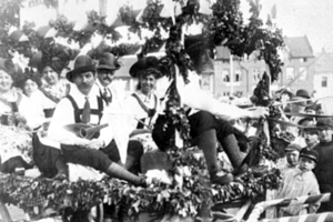 Eine Gruppe von Menschen in traditioneller bayrischer Tracht fährt auf einem mit Laub geschmückten Festwagen in einem Umzug, einige halten Musikinstrumente, mit Gebäuden und einem klaren Himmel im Hintergrund, dargestellt in Schwarz-Weiß.