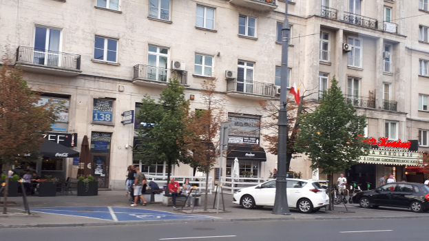Stadtstraße mit parkenden Autos, Bäume am Straßenrand, ein Gebäude mit Fenstern im Hintergrund, Fußgänger, die gehen und auf Bänken sitzen, Informationsschilder und ein Fahnenmast.