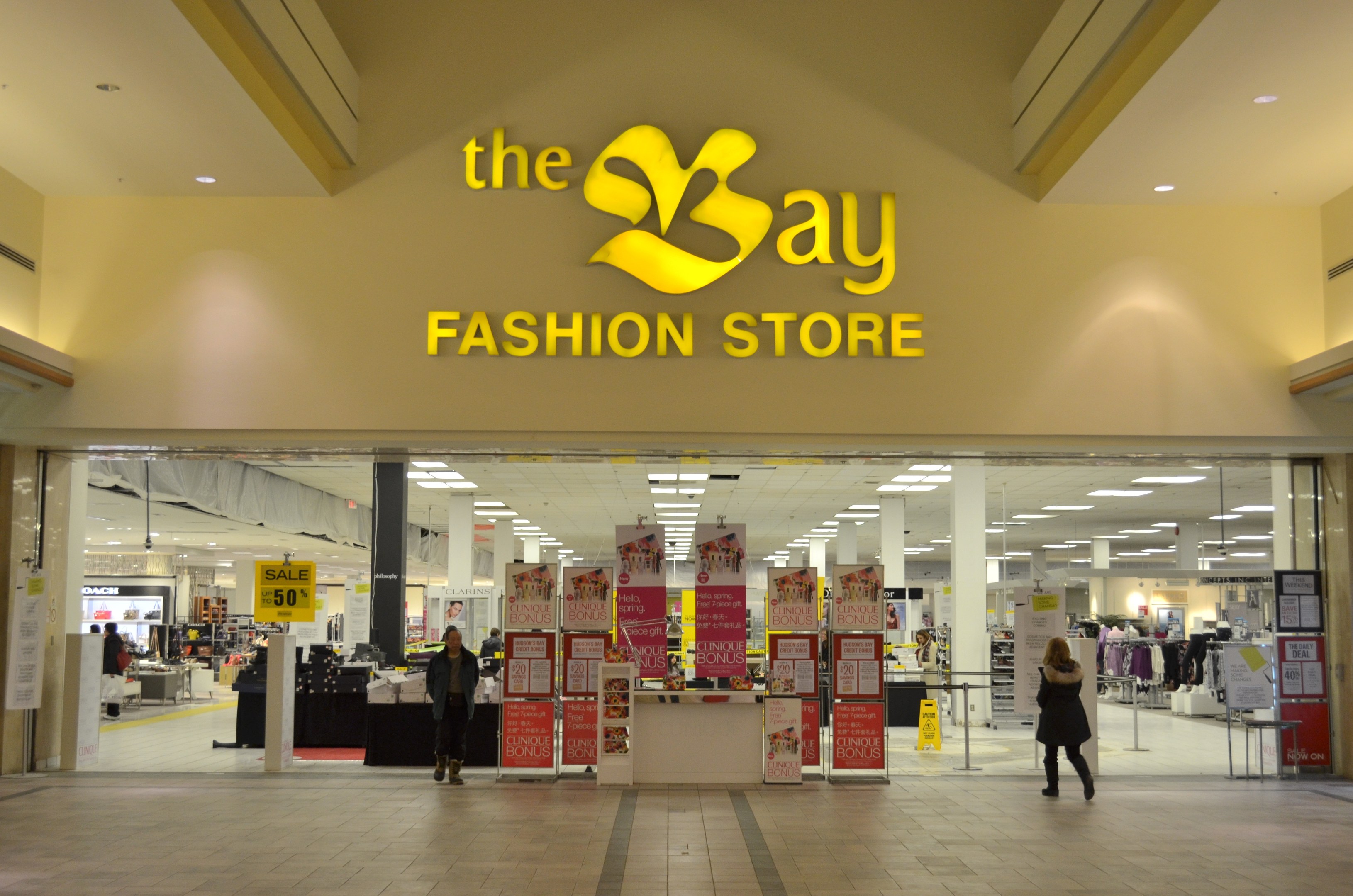 Außenansicht eines Bay Fashion Stores in einem Einkaufszentrum, zeigt Menschen, die gehen, Texttafeln an den Wänden, Säulen, Deckenleuchten und Hintergrundobjekte.
