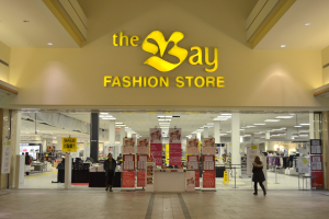 Außenansicht eines Bay Fashion Stores in einem Einkaufszentrum, zeigt Menschen, die gehen, Texttafeln an den Wänden, Säulen, Deckenleuchten und Hintergrundobjekte.