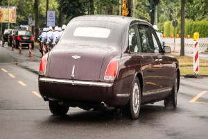 Ein braunes Auto fährt auf einer Straße neben einer Ampel, umgeben von Menschen in Helmen, Verkehrskegeln, Schildern, Polen, Verkehrszeichen, Fahnen, Pflanzen, Bäumen und Gras.
