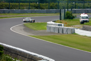 Ein Rennwagen fährt eine Strecke herunter mit einem Polizeiwagen im Hintergrund, umgeben von Grünfläche und Absperrung, mit Text unten auf dem Bild.