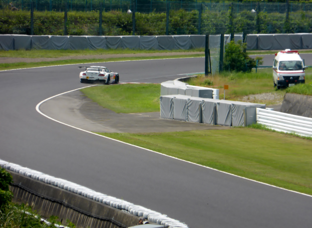 Ein Rennwagen fährt eine Strecke herunter mit einem Polizeiwagen im Hintergrund, umgeben von Grünfläche und Absperrung, mit Text unten auf dem Bild.