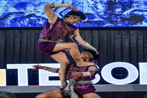 Zwei Performer in purpurfarbenen Kleidern führen einen akrobatischen Hebemanöver auf der Bühne vor einem Publikum aus, mit schwarzem Vorhang und Bildschirmhintergrund.