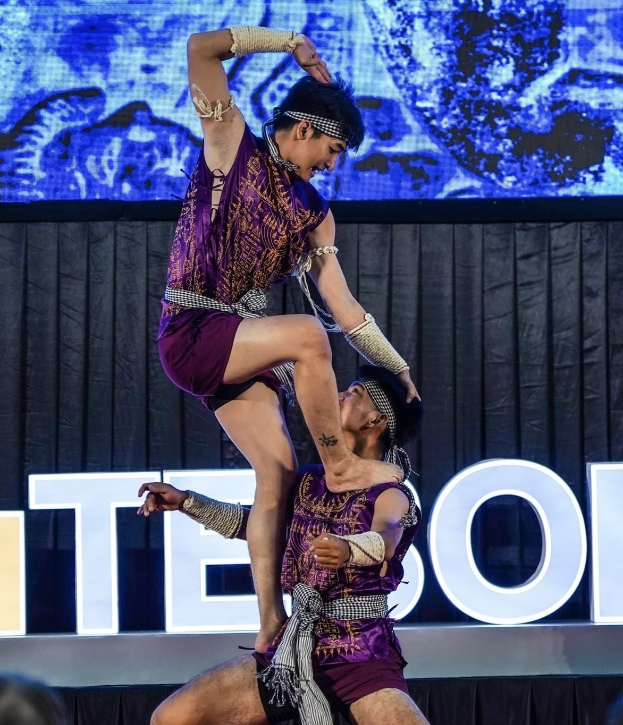 Zwei Performer in purpurfarbenen Kleidern führen einen akrobatischen Hebemanöver auf der Bühne vor einem Publikum aus, mit schwarzem Vorhang und Bildschirmhintergrund.