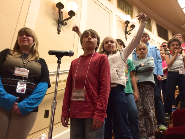 Eine Gruppe von Kindern singt an einem Mikrofon mit einer Frau daneben, Stühle rechts und beleuchtete Wände im Hintergrund.