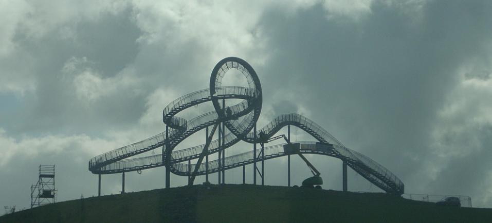 Achterbahn auf einem Hügel mit bewölktem Himmel, umgeben von Stützpfählen und Fahrgeschäfts-Infrastruktur.