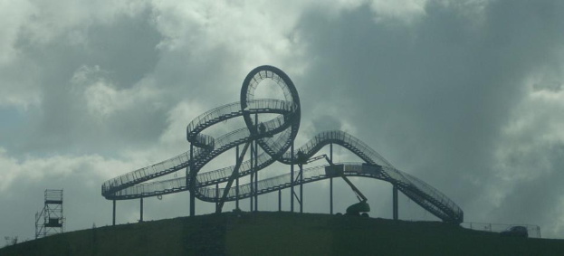 Achterbahn auf einem Hügel mit bewölktem Himmel, umgeben von Stützpfählen und Fahrgeschäfts-Infrastruktur.