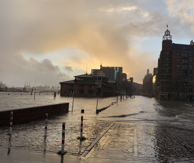 Überschwemmte Straße in Hamburg, Deutschland, mit Wasser auf der Straße, Pfählen, Schildern, Gebäuden mit Fenstern, einer Brücke und einem bewölkten Himmel im Hintergrund.