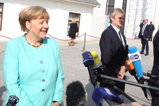 Bundeskanzlerin Angela Merkel beim Sprechen mit Reportern vor einem Gebäude, umgeben von Menschen mit Mikrofonen und einer Seilabsperrung unter einem bewölkten Himmel.