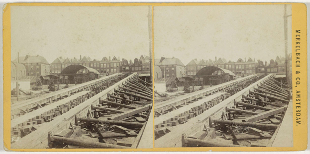 Schwarz-weiß-Foto einer Zugstrecke mit Gebäuden im Hintergrund, Text 'Merkelbach & Co, Co. Amsterdam' auf der rechten Seite und verstreute hölzerne Gegenstände im Vordergrund.
