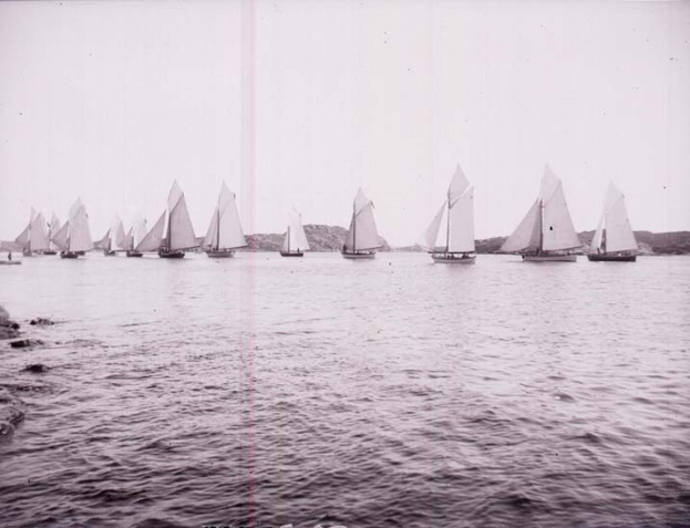 Ein Schwarz-Weiß-Foto mehrerer Segelboote auf dem Wasser mit Hügeln im Hintergrund und einem klaren Himmel darüber, mit Text unten, der darauf hinweist, dass es ein Foto des New York Yacht Club ist.