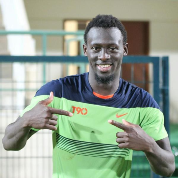 Ein Mann in einem grün-blauen Hemd, der lachend in die Kamera zeigt und im Hintergrund eine Reling, eine Wand und eine Tür zu sehen sind.