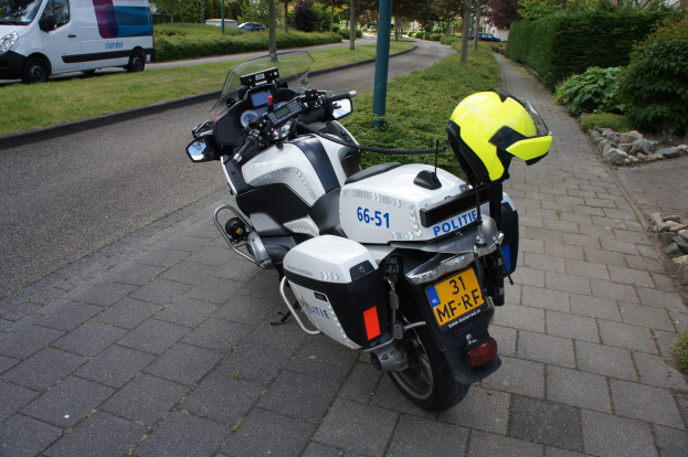 Eine Polizeimotorrad auf dem Seitenstreifen geparkt mit einem Helm darauf, umgeben von Bäumen, Polen, Gebäuden, Pflanzen, Gras, Steinen und anderen Fahrzeugen.