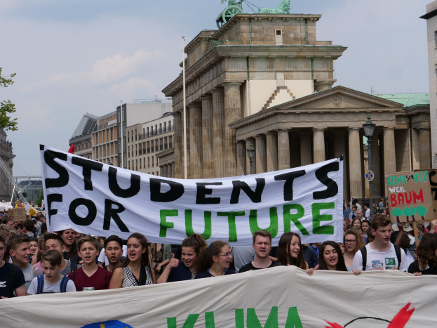 Gruppe von Schülern, die in Berlin mit einem bunt bemalten 'Schüler für die Zukunft'-Schild marschieren, vor dem Hintergrund von Gebäuden, Bäumen und Himmel.