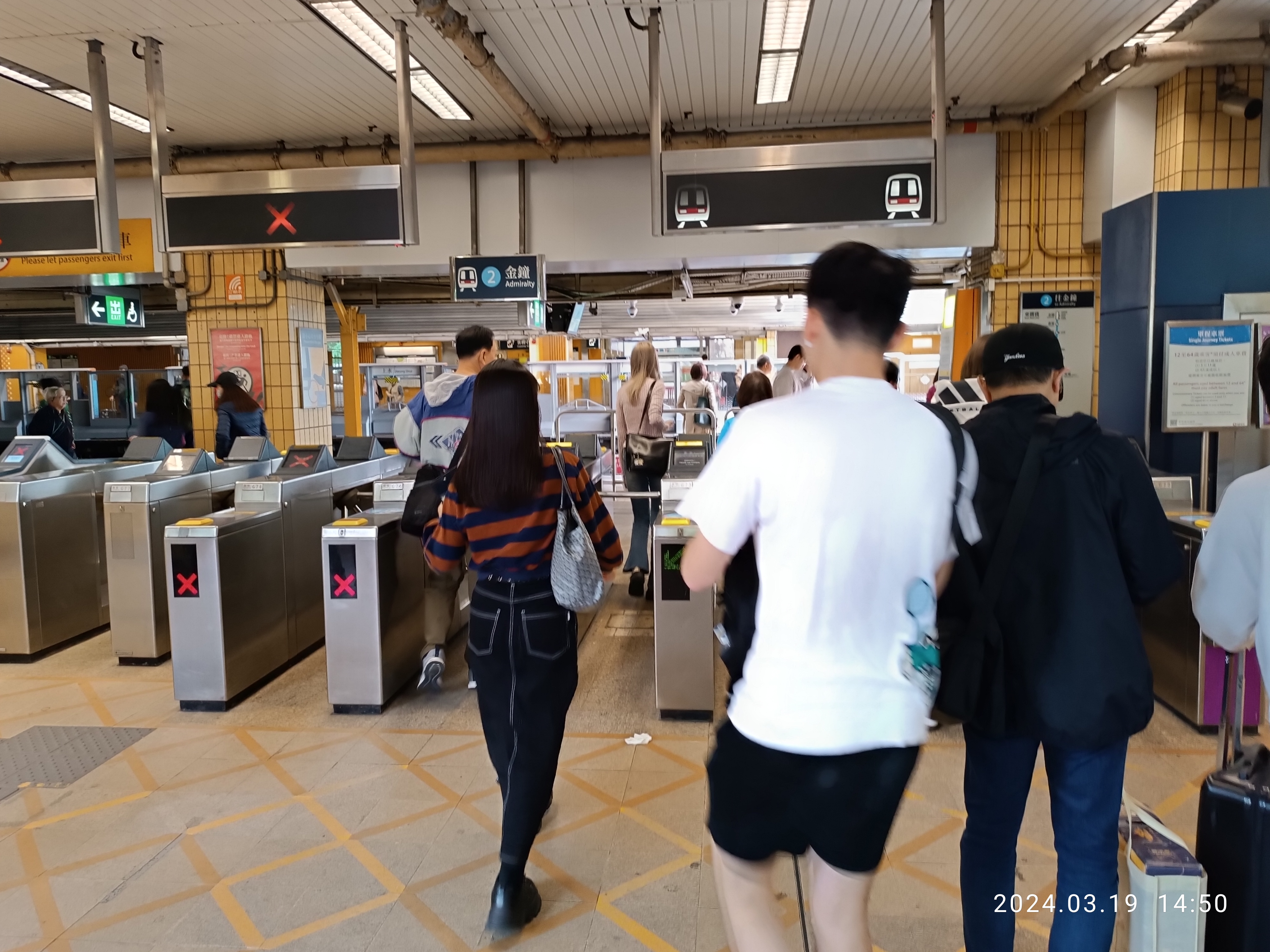 Eine Gruppe von Menschen geht durch einen Bahnhof, einige tragen Taschen, mit Metalldetektoren, Schildern, Metallstangen und Deckenleuchten sowie einem Wasserzeichen im rechten unteren Eck.