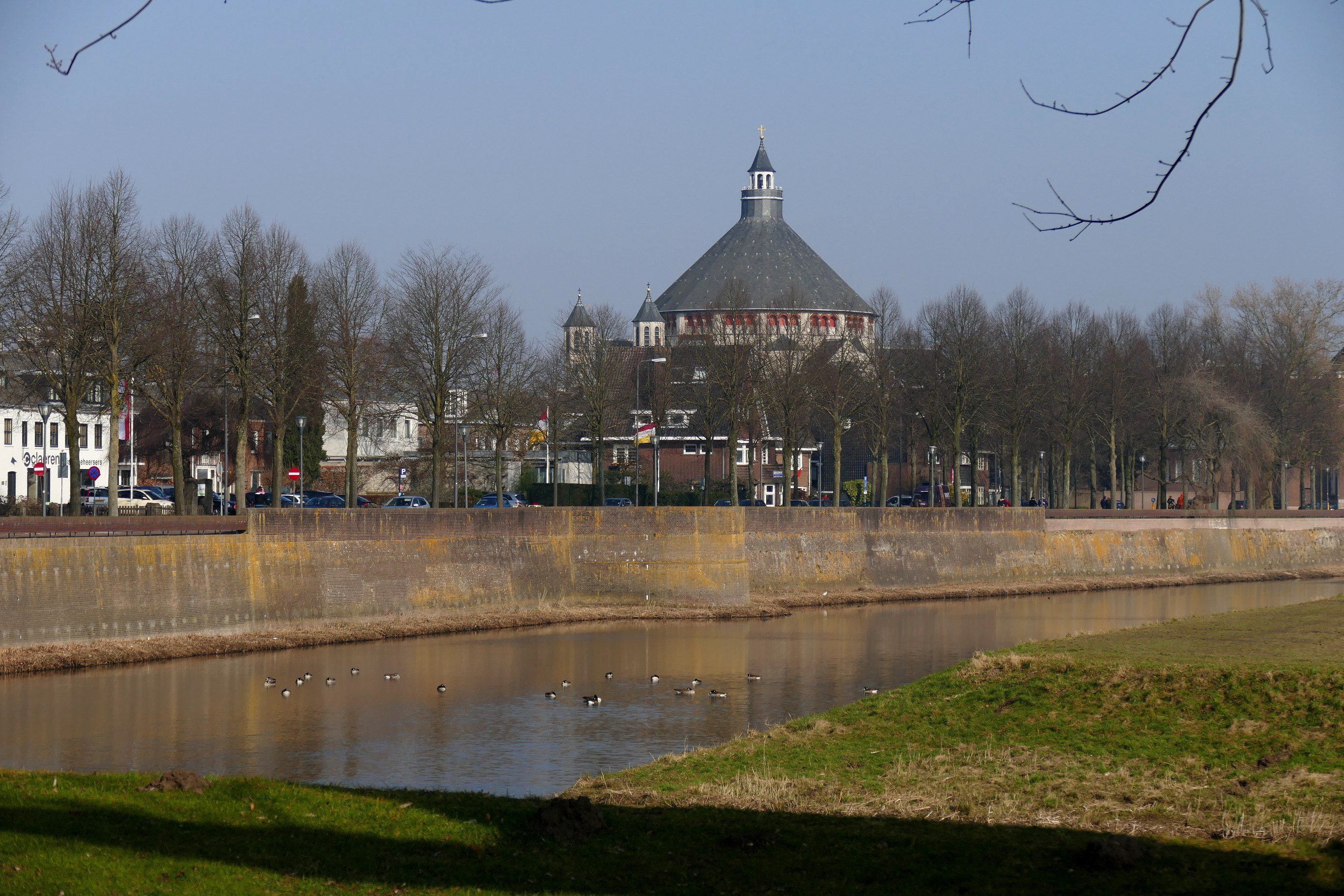 Ein Fluss mit schwimmenden Enten, gesäumt von grünem Gras und Bäumen, mit Gebäuden, Fahrzeugen, Pfählen, Flaggen und einem klaren blauen Himmel im Hintergrund.