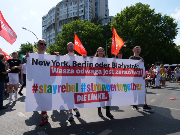 Eine Gruppe von Menschen, die eine Straße in Berlin, Deutschland, entlanggehen und ein Banner mit der Aufschrift "Stay Rebel Stronger Together" halten, mit Bäumen, Laternenpfählen und Gebäuden, die die Straße säumen, unter einem bewölkten Himmel.