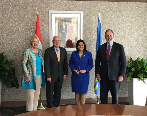 Eine Gruppe von Menschen, die um einen Tisch mit Papieren stehen und lächeln, umgeben von Fahnen, Topfpflanzen und einem Rahmenfoto an der Wand, was das Unterzeichnen eines U.S.-Kanada-Memorandums angibt.