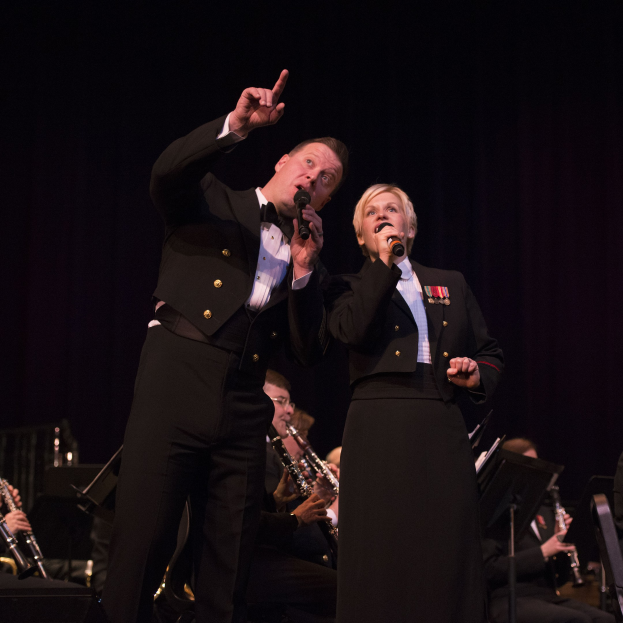 Ein Mann und eine Frau in Fracks, die Mikrofone halten, auf einer Bühne mit einem sitzenden Orchester dahinter in einer dunklen Umgebung.
