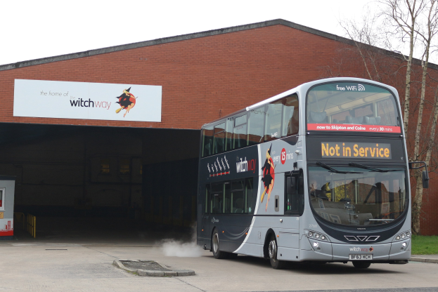 Doppeldeckerbus mit Rauchwolke an einem Gebäude vorbeifahrend, mit Gras, Bäumen und einem klaren Himmel im Hintergrund und einem sichtbaren Fahrzeugkennzeichen am Bus.