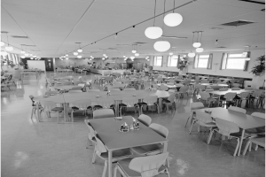 Ein geräumiger Speisesaal mit Reihen von Tischen und Stühlen, verstreuten Flaschen und Gegenständen auf den Tischen, Deckenlampen, Fenstern auf der rechten Seite und Topfpflanzen überall im Raum.