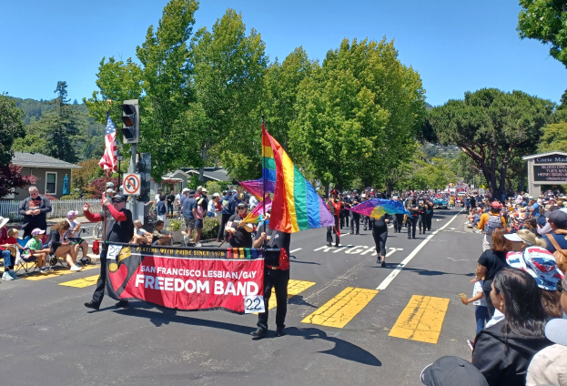 Eine Gruppe von Menschen, die auf der Straße einer Parade marschieren, einige mit Schirmen, andere sitzend oder stehend, mit einer Regenbogenflagge, einem Verkehrssignal, einem Schild, Bäumen, Gebäuden und dem Himmel im Hintergrund.