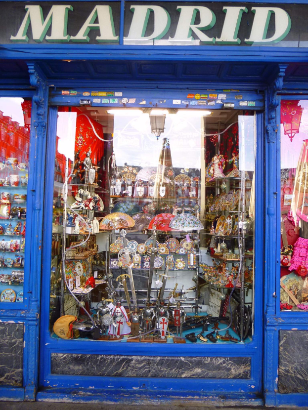 Ein Schaufenster mit einer blauen Tür und einem "Madrid"-Schild, das verschiedene Gegenstände zeigt, die wie eine belebte Stadtlandschaft angeordnet sind.