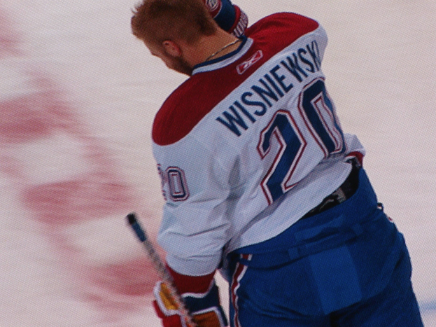 Ein Eishockeyspieler in einem "Wisniewski"-Trikot steht auf dem Eis, hält einen Eishockeyschläger und hat einen konzentrierten Gesichtsausdruck.