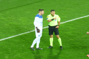 Ein Schiedsrichter in gelbem T-Shirt und schwarzen Shorts spricht mit einem Fötballspieler in weiß-blauer Uniform auf dem Feld, beide tragen Turnschuhe.