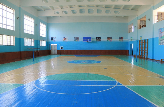 Großer Sportraum mit blau-weißem Boden, einem Basketballfeld, einem Netz mit Stangen, einer Tür, Fenstern, Wandplakaten, einem Geländer und einem Dach.