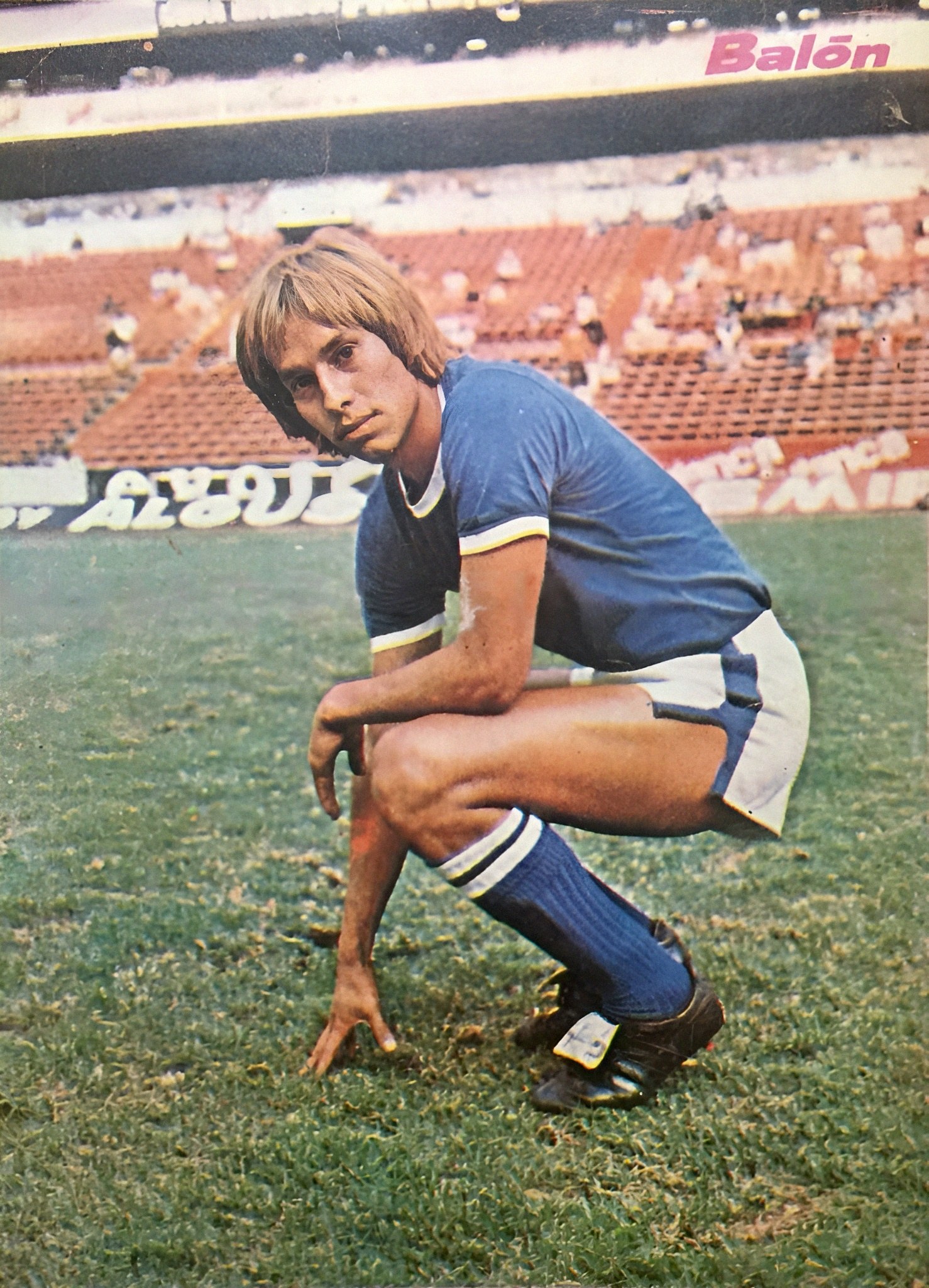 Ein Mann kniet auf einem Fußballfeld umgeben von Menschen mit Schildern, trägt Sportschuhe.