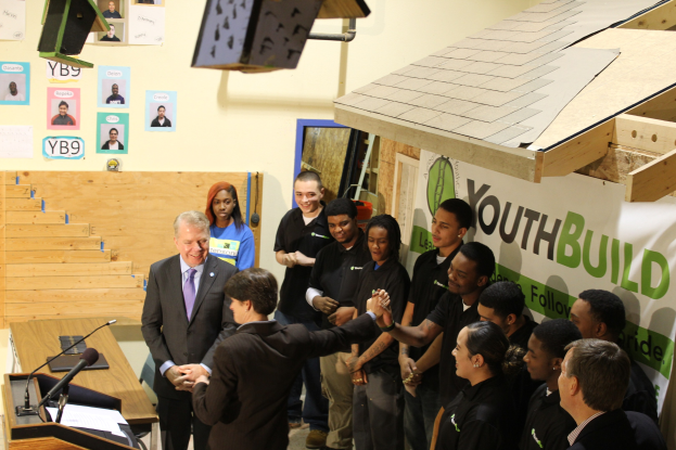 Gruppe von Menschen, die sich die Hände schütteln, um einen Podium mit Mikrofonen und Papieren auf einem Tisch, vor einem Publikum, mit Plakaten an der Wand und Gegenständen auf dem Dach und der rechten Seite.