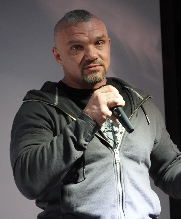 Ein Mann in einer schwarzen Kapuzenjacke und einem schwarzen Anzug mit einem weißen Hemd, der ein Mikrofon hält, steht selbstbewusst vor einer Wand und scheint ein Komiker mit zurückgekämmtem Haar zu sein.