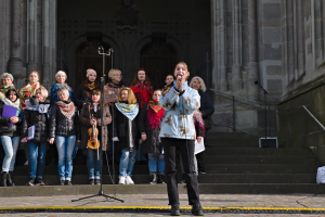 Eine Gruppe von Menschen, einige mit Musikinstrumenten, steht vor einem Gebäude mit Treppen und Geländern, während eine Frau in der Mitte in ein Mikrofon singt, umgeben von Mikrofonstativen.