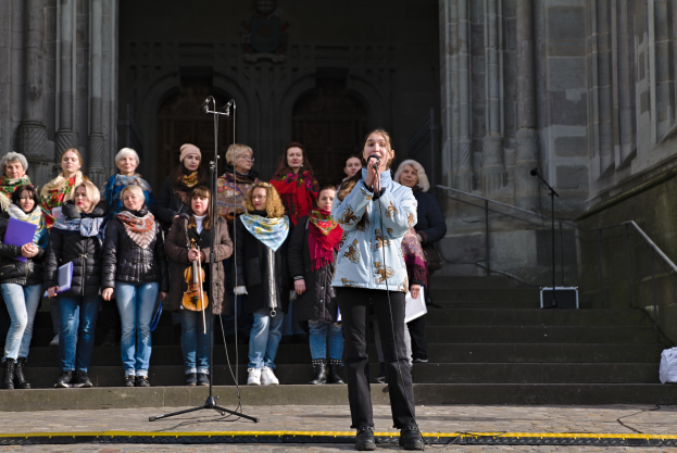 Eine Gruppe von Menschen, einige mit Musikinstrumenten, steht vor einem Gebäude mit Treppen und Geländern, während eine Frau in der Mitte in ein Mikrofon singt, umgeben von Mikrofonstativen.