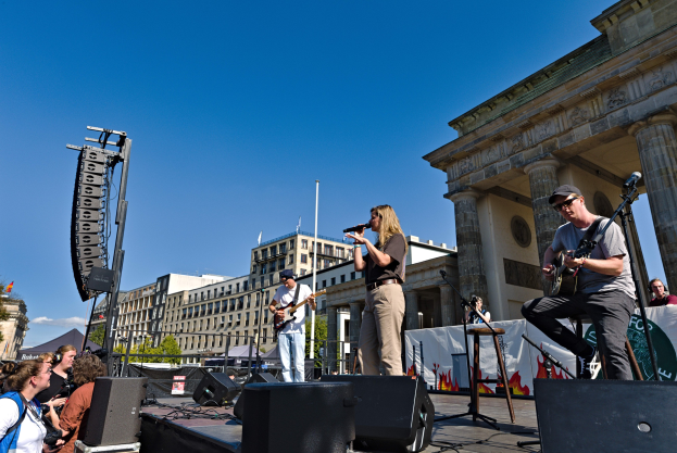 Eine Gruppe von Menschen auf einer Bühne vor dem Brandenburger Tor in Berlin, Deutschland, die Musikinstrumente spielen und in Mikrofone singen, mit Lautsprechern und anderen Gegenständen drumherum und Gebäuden, Bäumen, Pfählen, Flaggen und einem klaren blauen Himmel im Hintergrund.