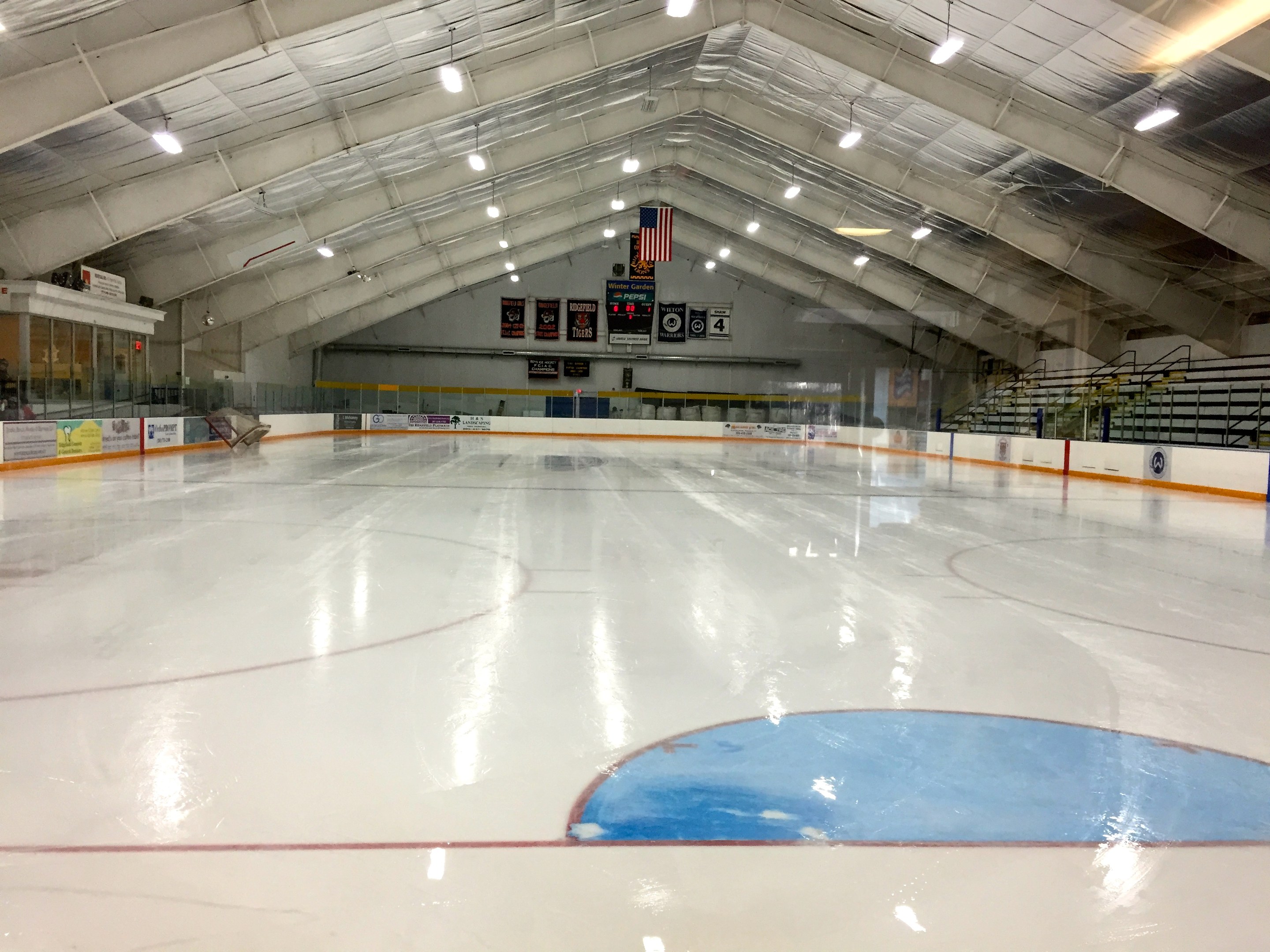 Große Indoor-Eisfläche mit Deckenbeleuchtung, Texttafeln, einer Wand, Treppen mit Geländern und einigen Menschen, was auf eine Sportanlage hinweist, wahrscheinlich eine Eishockeyhalle.