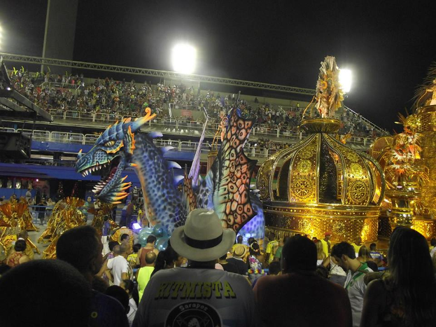 Eine große Menschenmenge, die um einen bunt geschmückten, hell erleuchteten Karnevalswagen steht, mit einem Stadion voller Menschen im Hintergrund und dem Himmel darüber.
