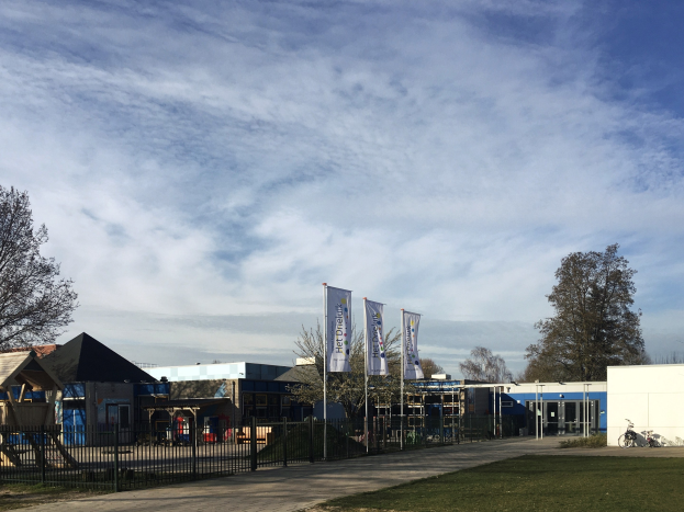 Großes Backsteinschulgebäude mit einem grasbewachsenen Bereich, der von einem Metallzaun umgeben ist, mit einem Weg, Bäumen, Fahnen, einem geparkten Fahrrad, nahen Häusern und einem bewölkten Himmel.