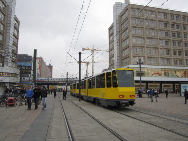 Eine gelbe Straßenbahn auf einer Stadtstraße mit Fußgängern in der Nähe, Fahrräder auf dem Gehweg abgestellt und Gebäude, Masten, Drähte, einen Kran und den Himmel im Hintergrund.
